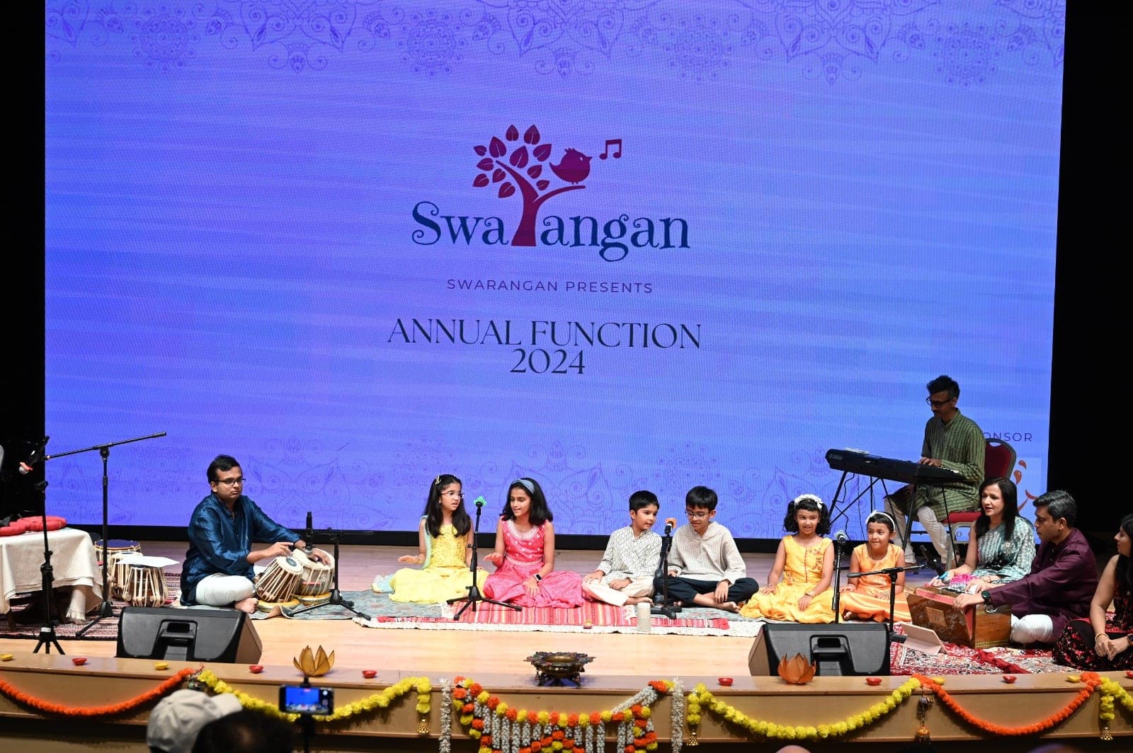 Young Swarangan students singing together on stage at the 2024 annual function, accompanied by tabla and harmonium